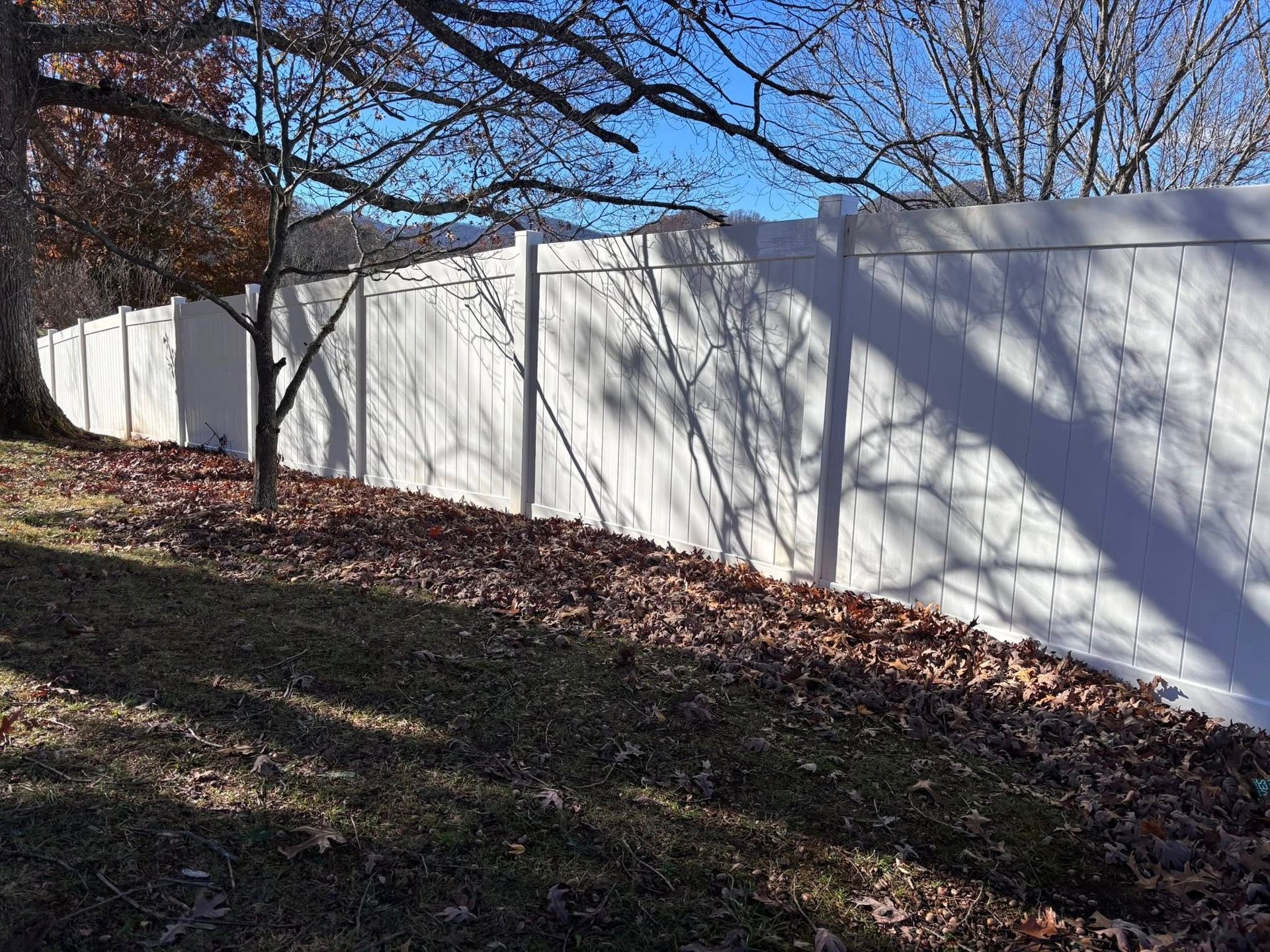 White wooden fence along a grassy, leaf-covered hillside under bare trees and blue sky