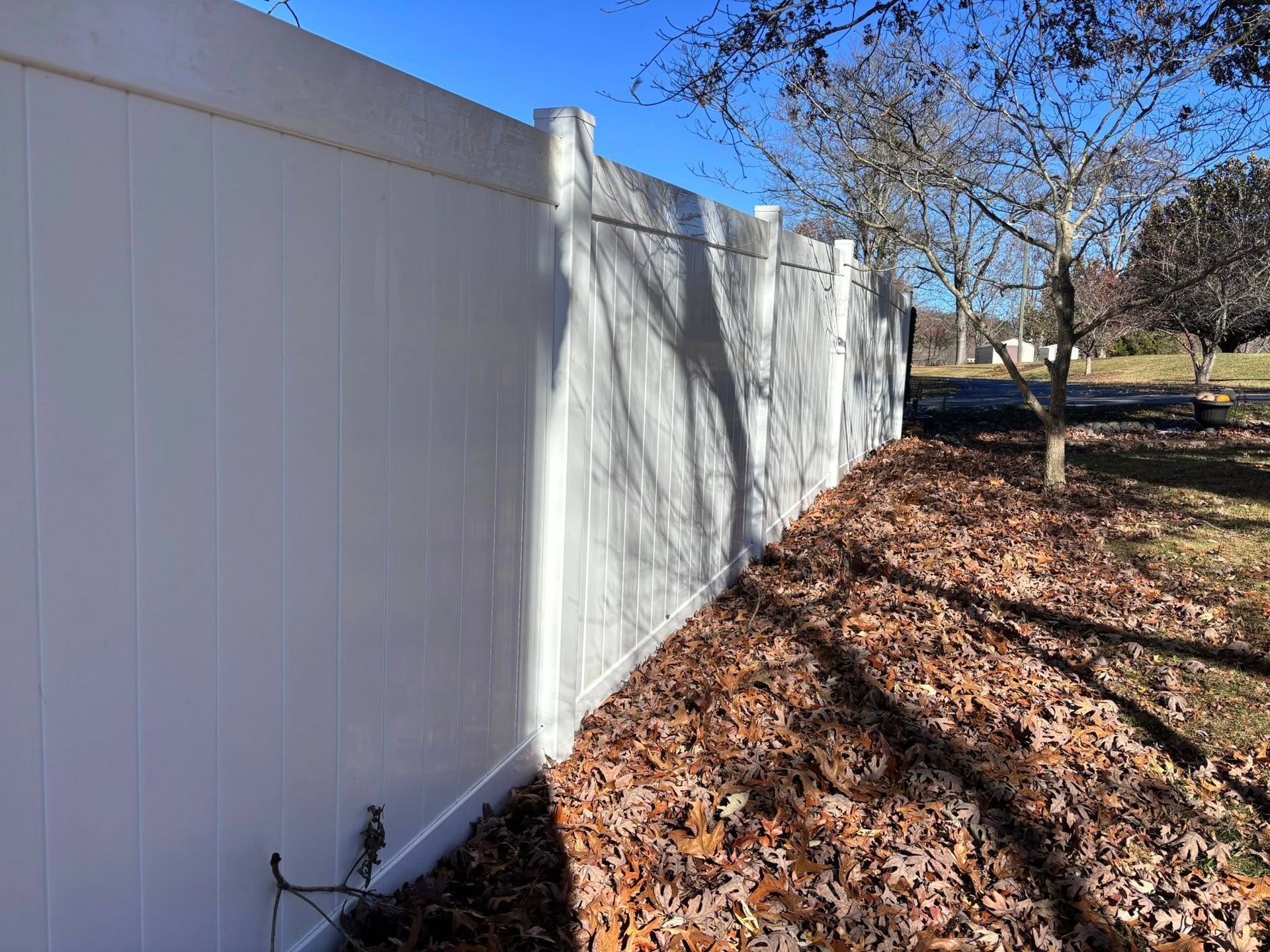 White vinyl fence along a leaf-covered yard with tree shadows on a sunny day