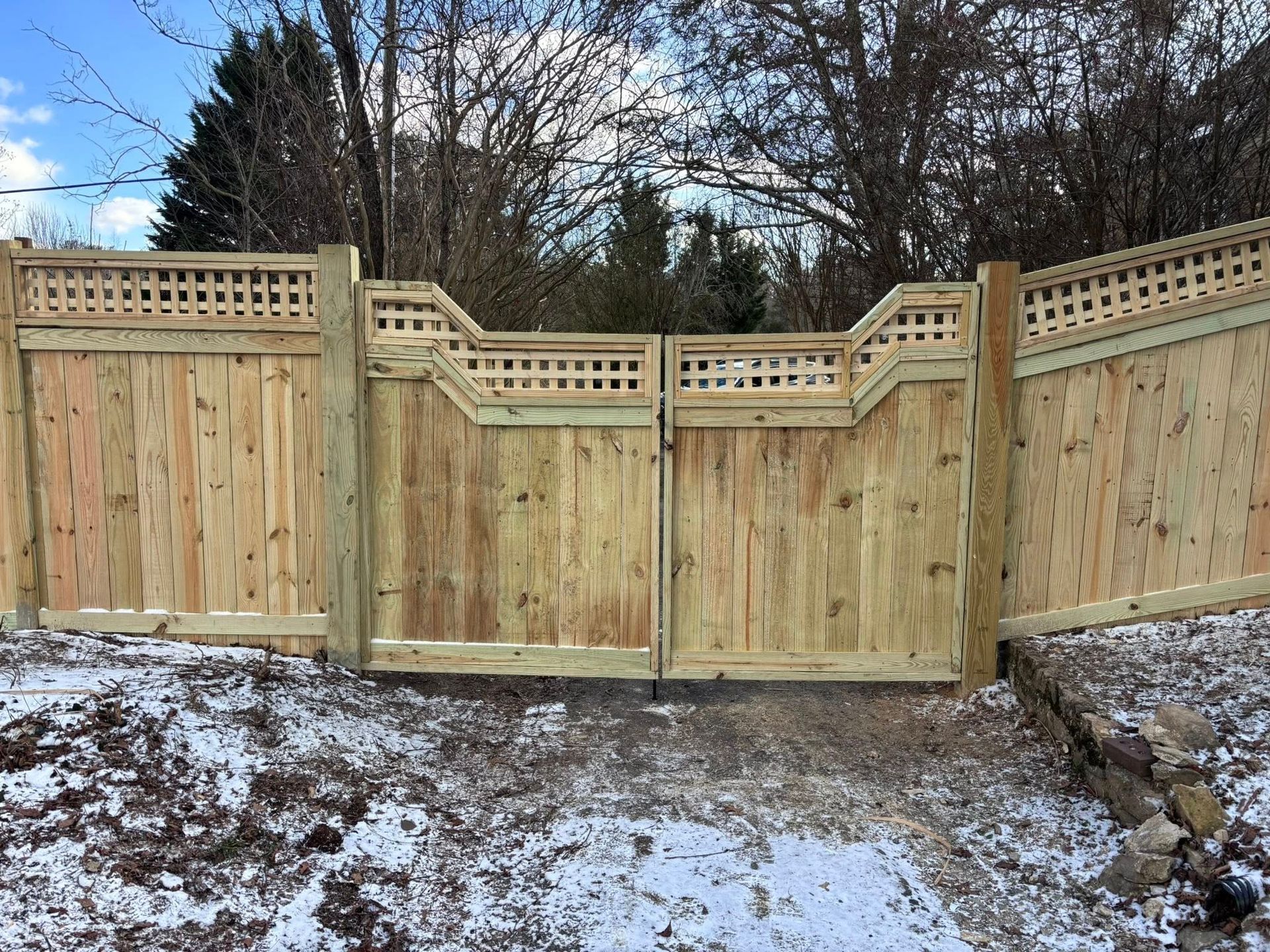 Wooden privacy fence with gates on a snowy yard at the edge of a wooded area