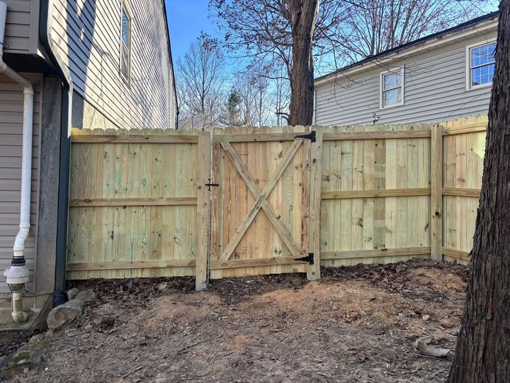 A wooden privacy fence with a central X-braced gate standing between two houses in a yard covered with brown leaves.
