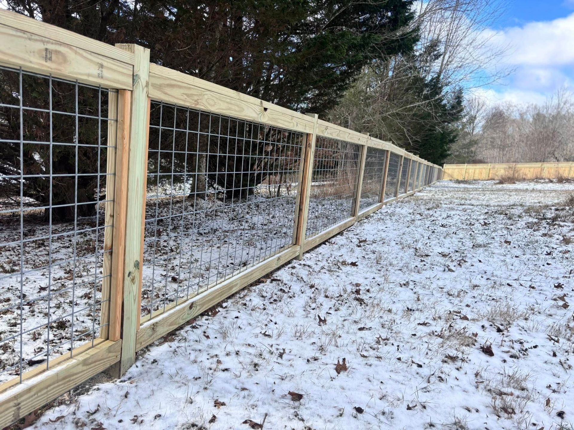 Wooden wire fence lining a snowy field beside trees and an open yard