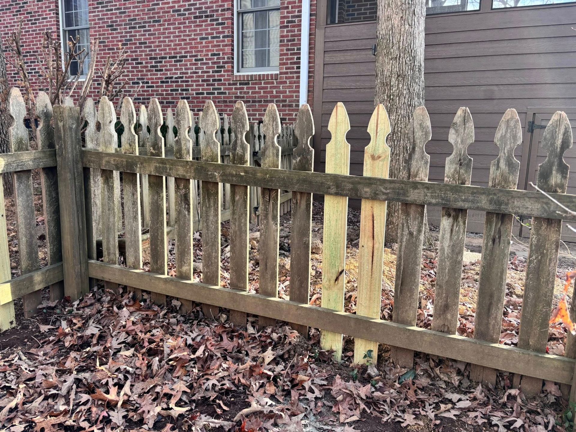 Weathered wooden picket fence beside a brick house, with fallen leaves on the ground.