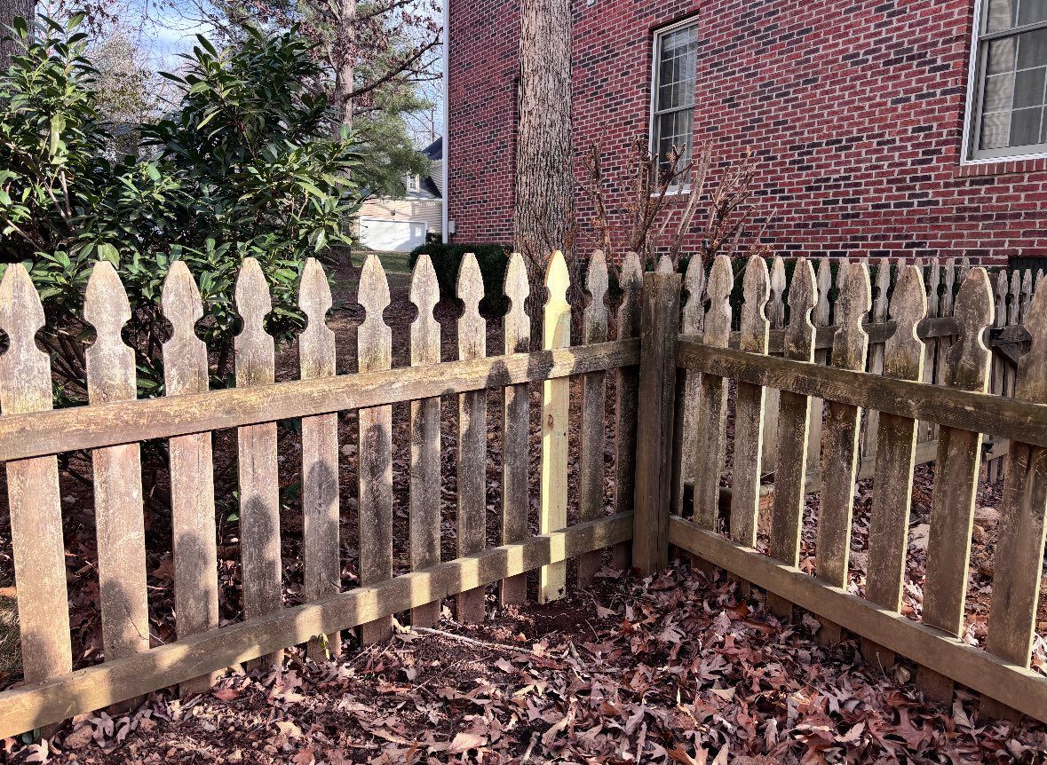 Wooden picket fence in a leaf-covered yard beside a red brick house, with shrubs behind it
