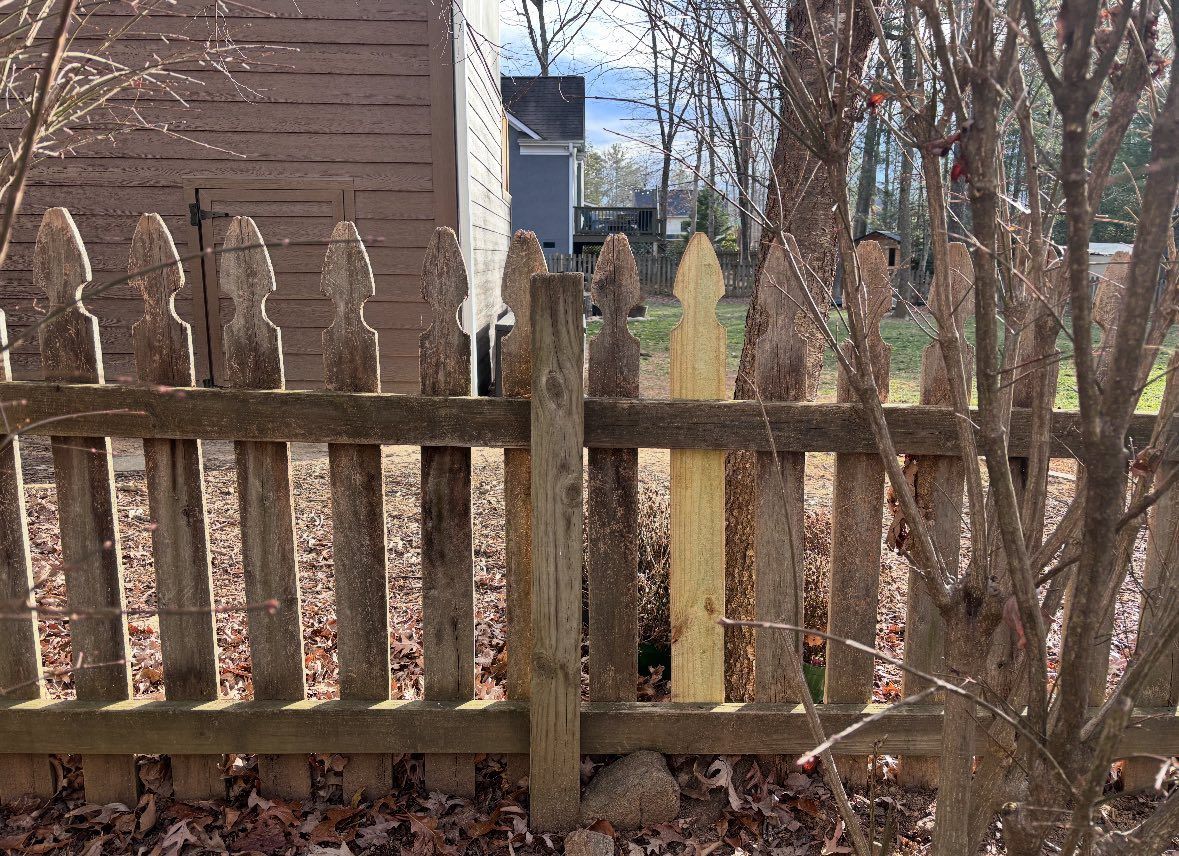 Weathered wooden fence with pointed slats in a leaf-covered yard beside a brick house