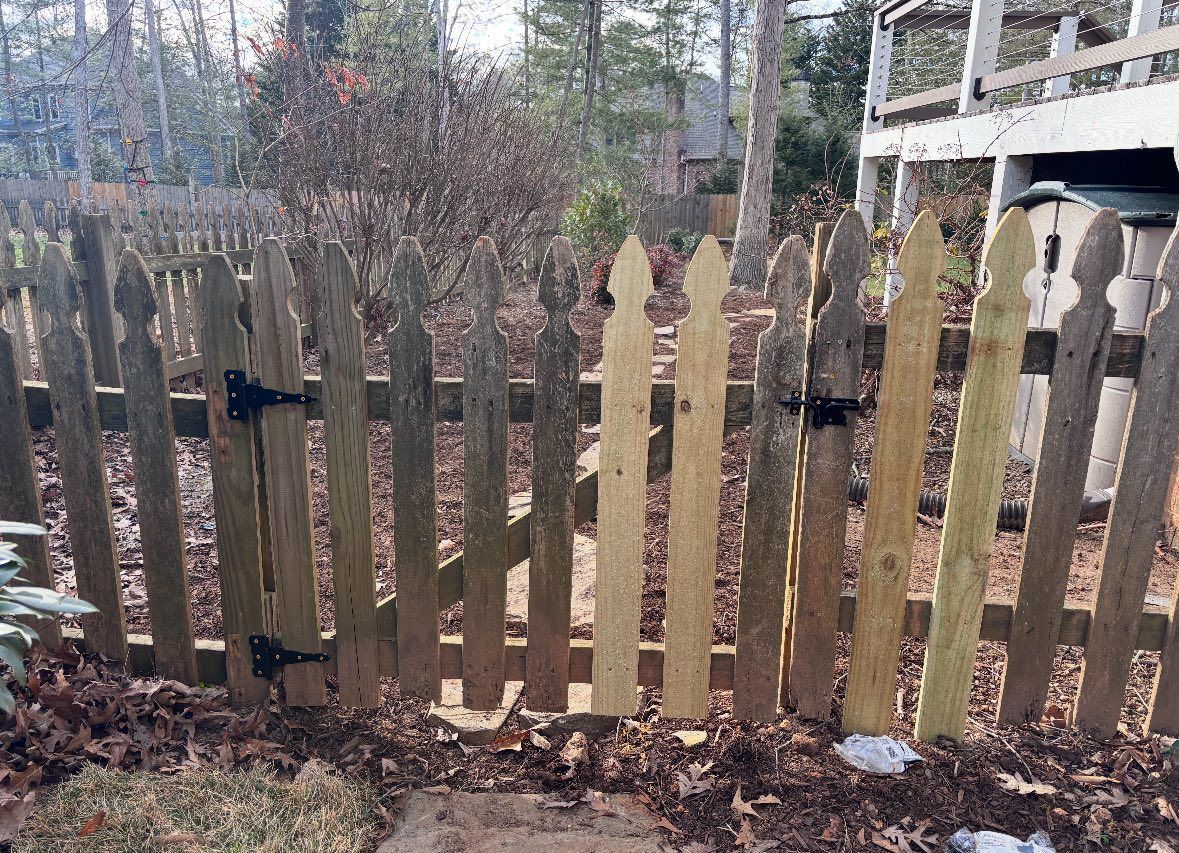 Weathered wooden picket fence in a leaf-covered yard, with trees and a carport in the background