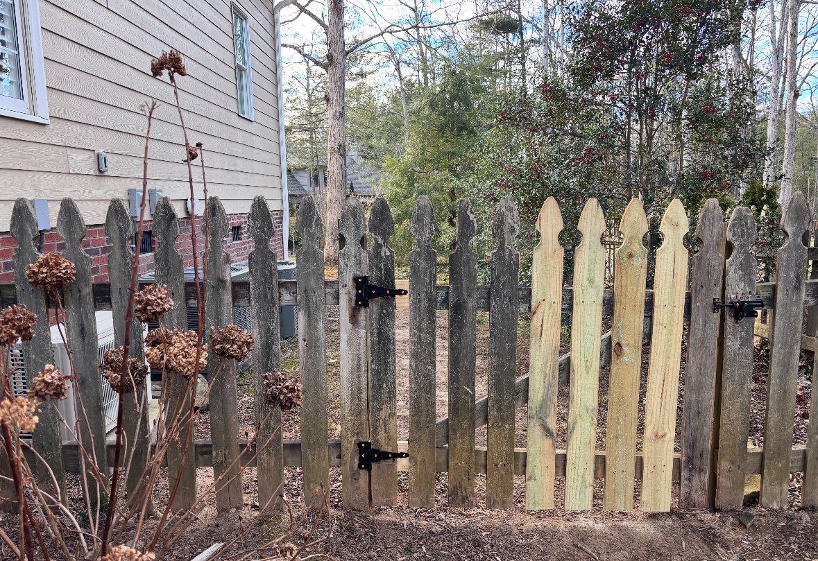 Weathered wooden picket fence in a yard beside a house, with bare trees and shrubs in the background