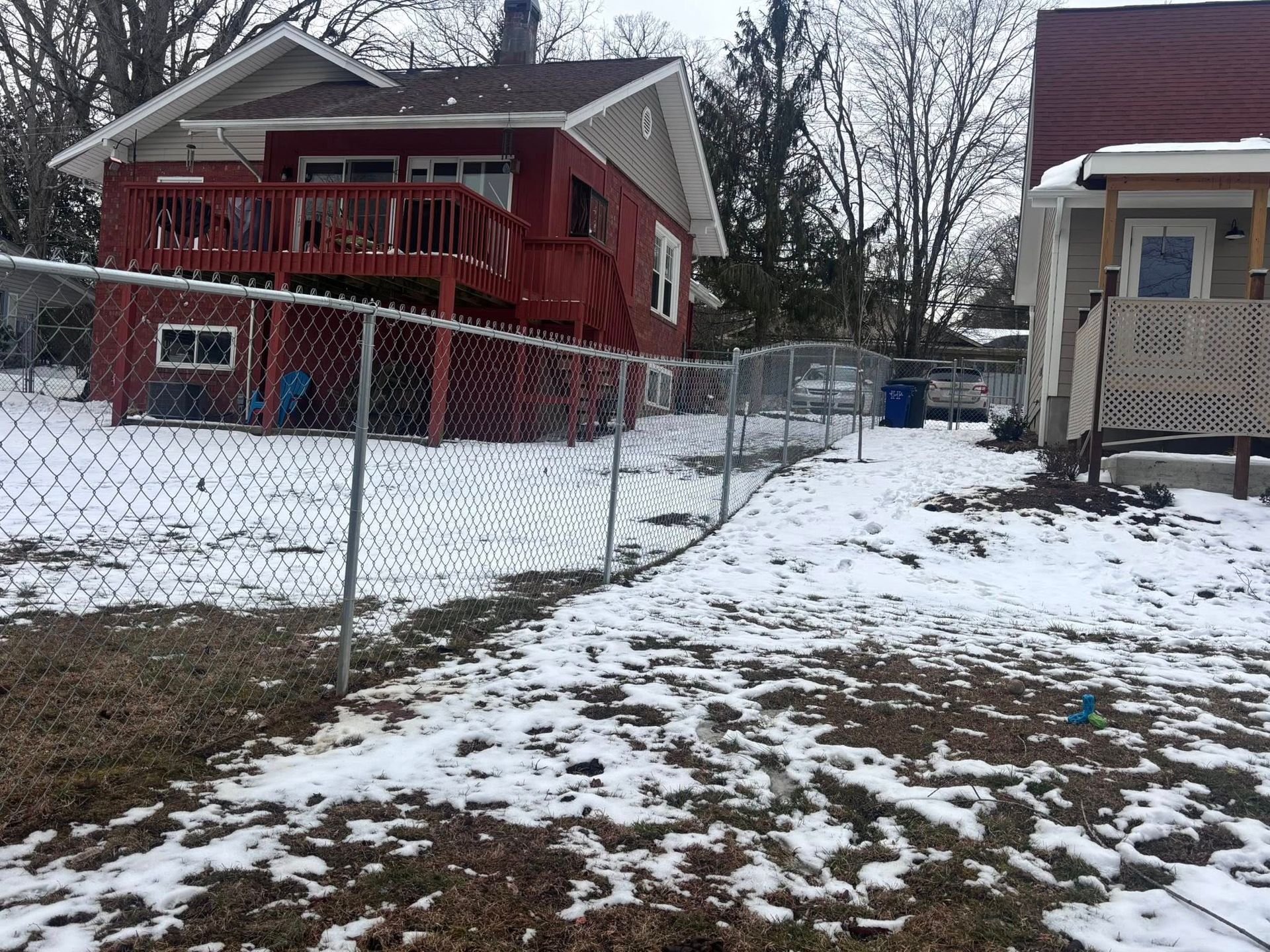 Snowy residential alley between a red house and a beige house behind a chain-link fence.