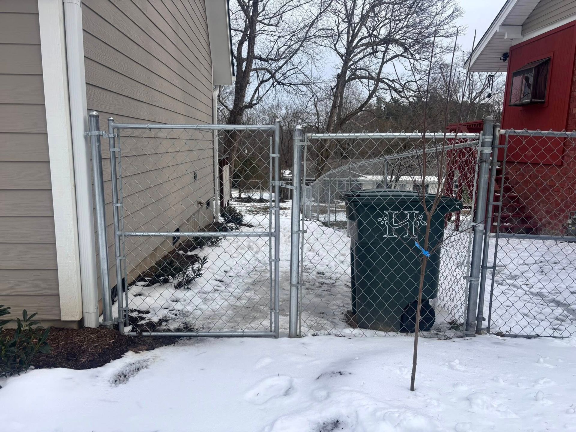 Snowy side yard with chain-link gate and fence beside a house and red building
