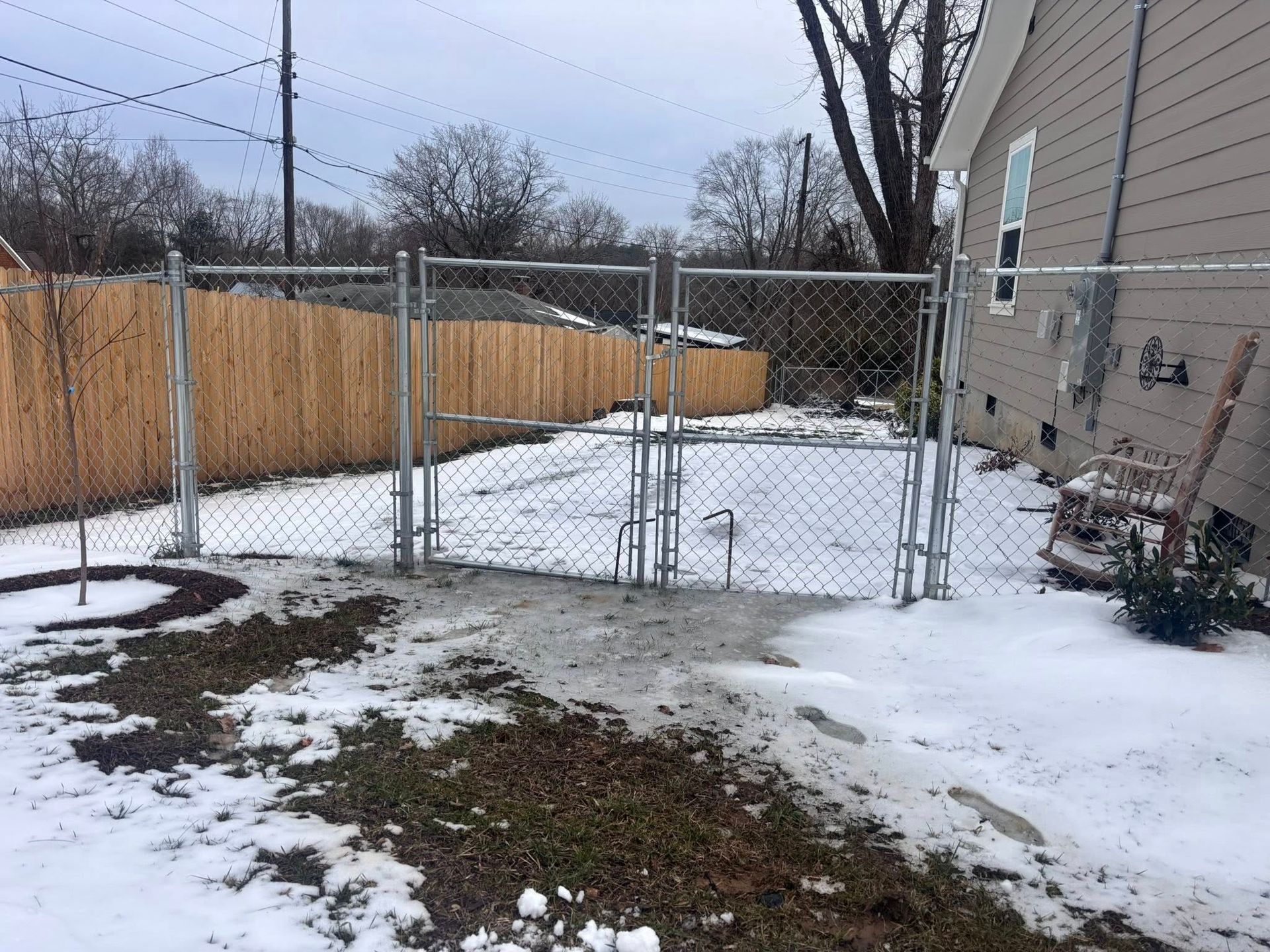 Snowy backyard with chain-link fence, beige house, wooden privacy fence, and bare trees.