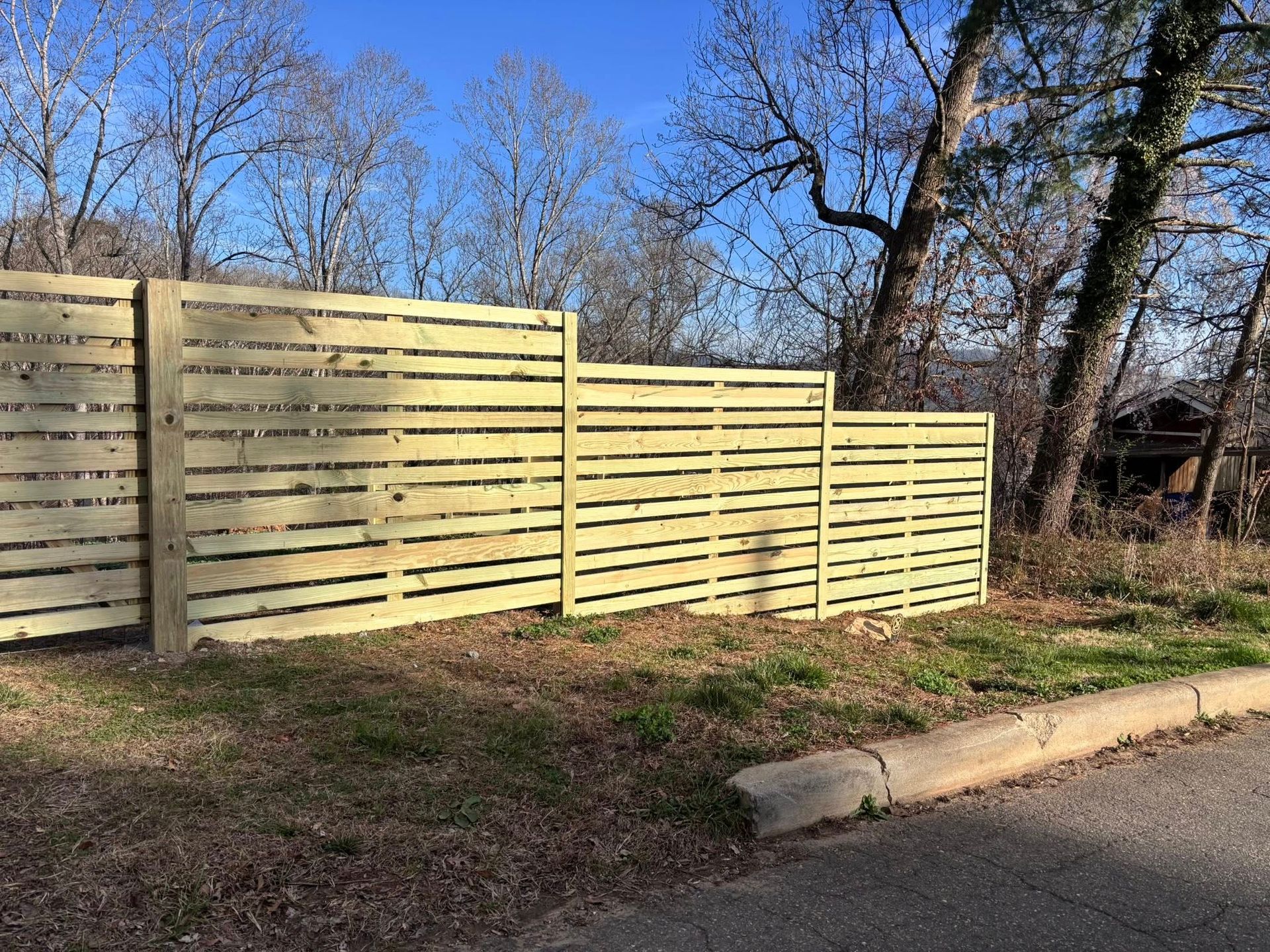 Wooden privacy fence along a roadside, with bare trees and a grassy verge on a clear day