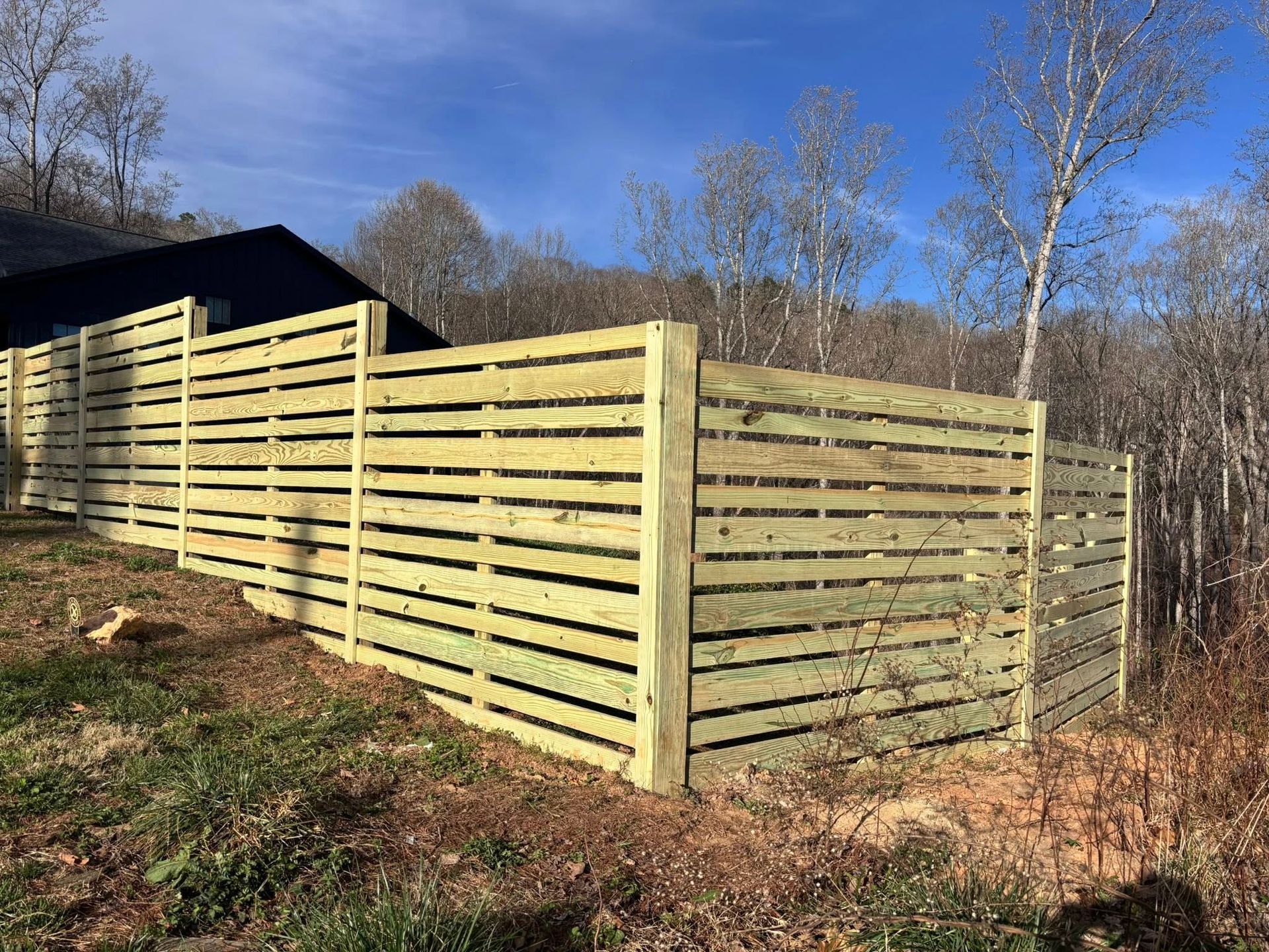Long wooden slat fence enclosing a yard beside a dark shed on a sunny hillside.