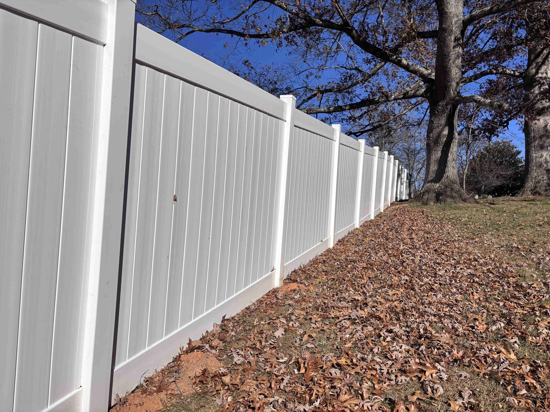 White vinyl fence lining a leaf-covered hillside under a blue sky with bare trees