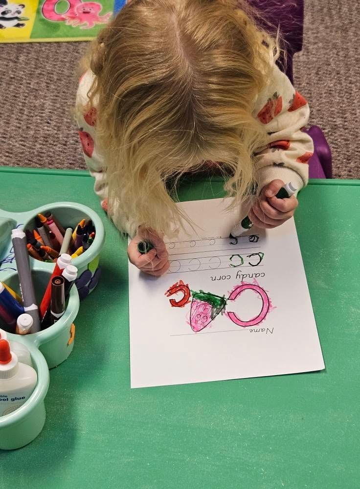 Child coloring a worksheet with markers at a green table.
