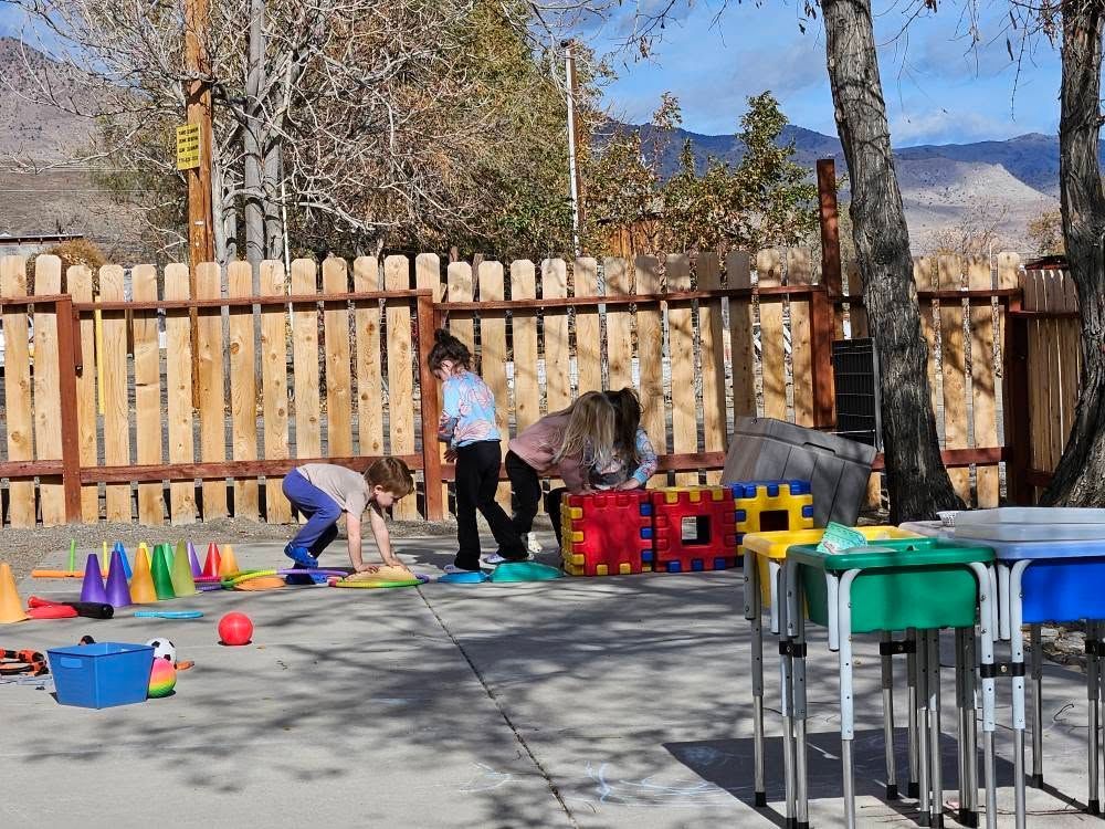 Children playing with colorful toys outdoors beside a wooden fence and tables, with mountains in the background