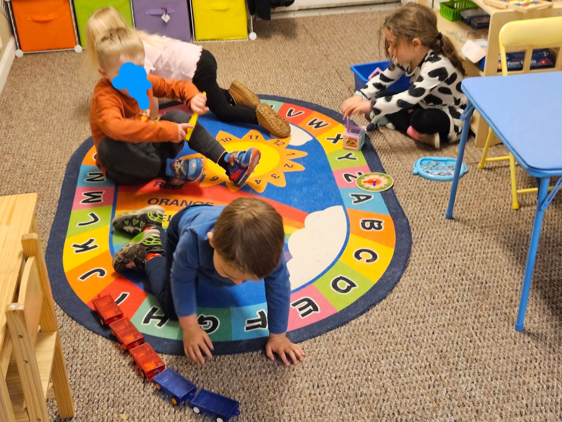 Children playing on a colorful circular classroom rug with toy vehicles and blocks.