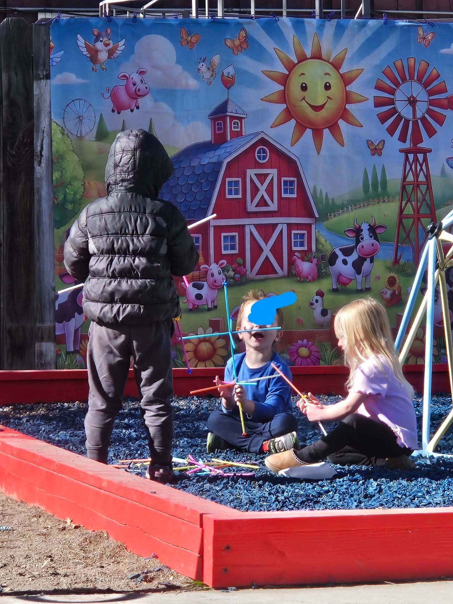 Children playing in a sandbox before a colorful farm mural, with a red playhouse and bright sun backdrop