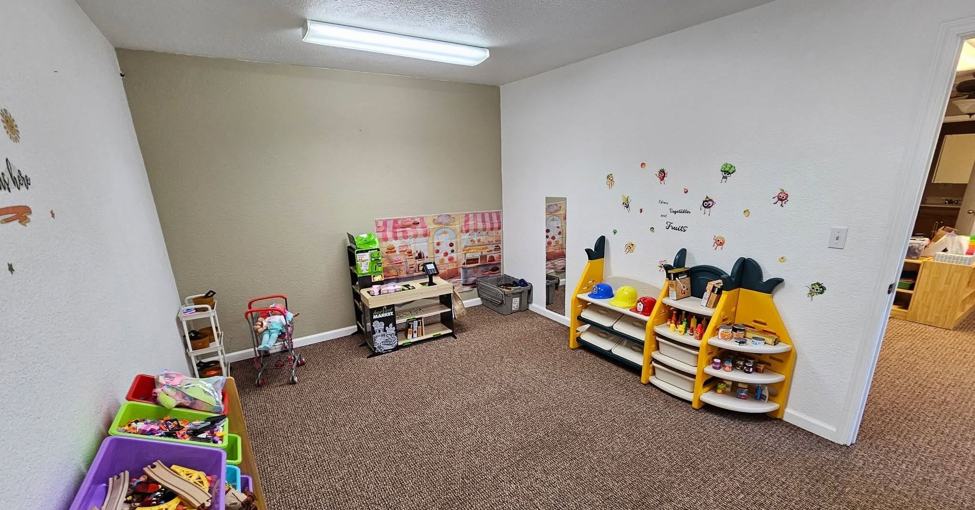 Small classroom play area with shelves of toys, books, and colorful bins on carpeted floor