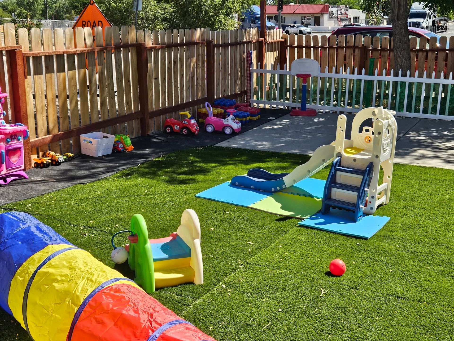 Backyard playground with colorful slide, toy cars, and bright turf beside a wooden fence