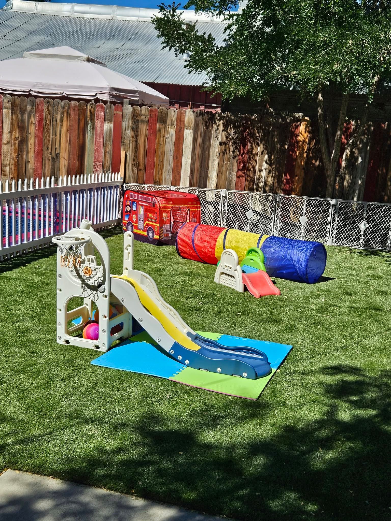 Backyard children’s play area with a small slide, tunnel, and colorful play structures on grass