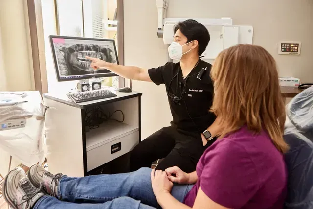 A dentist is talking to a patient in a dental chair while pointing at a computer screen.