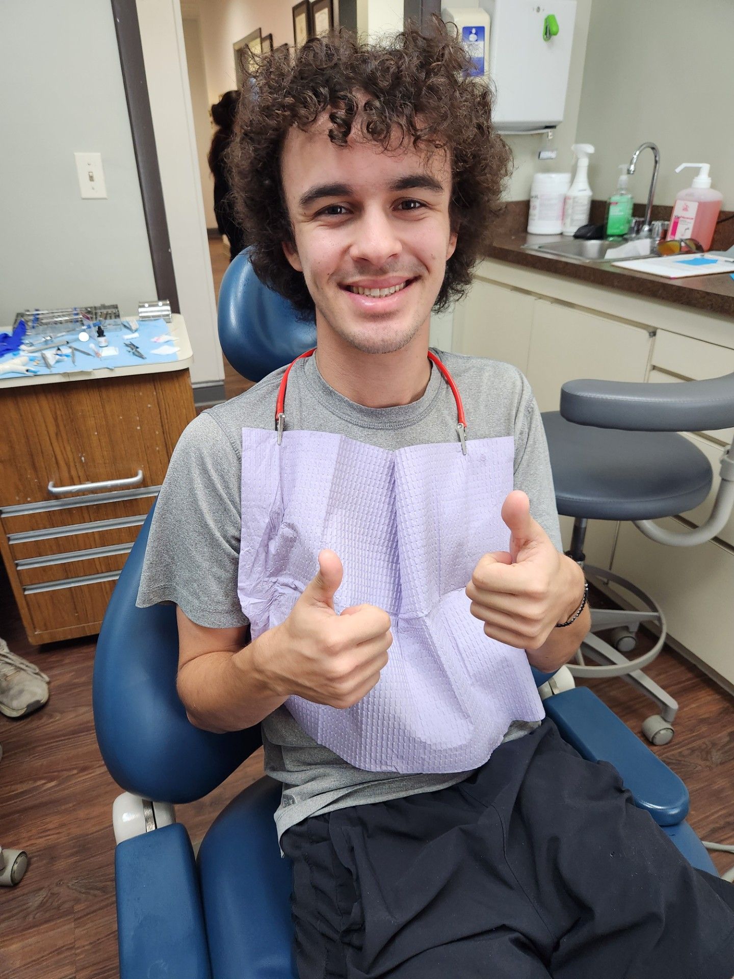 Un joven en el sillón del dentista, con el pulgar hacia arriba y un babero puesto, tiene el pelo rizado y sonríe.