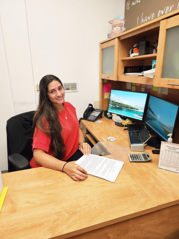 Mujer con cabello largo castaño, camisa roja, sentada en un escritorio con dos monitores, sonriendo.