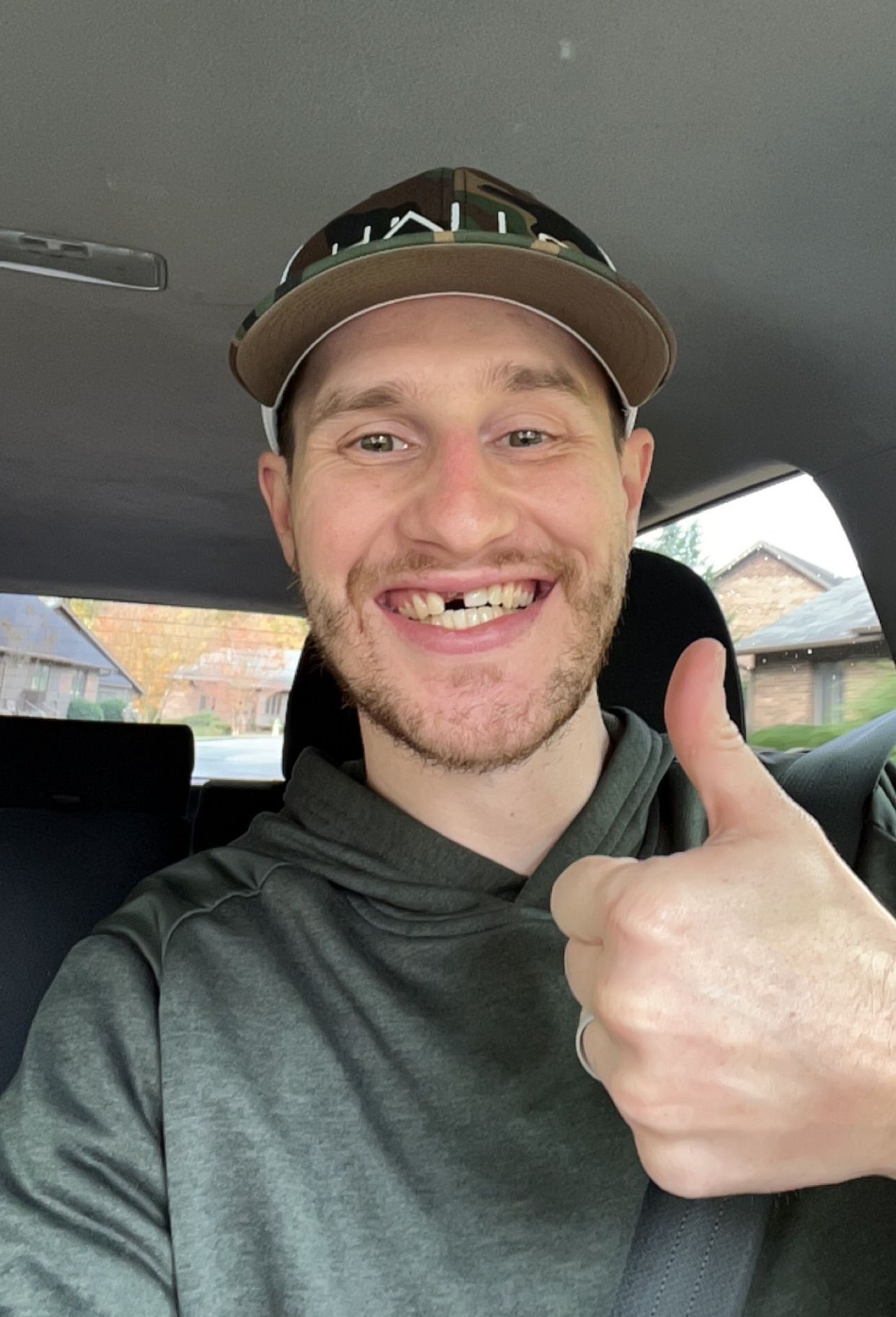 Hombre en coche hace el gesto de aprobación con el pulgar, sonriendo, lleva gorra y le falta un diente frontal.