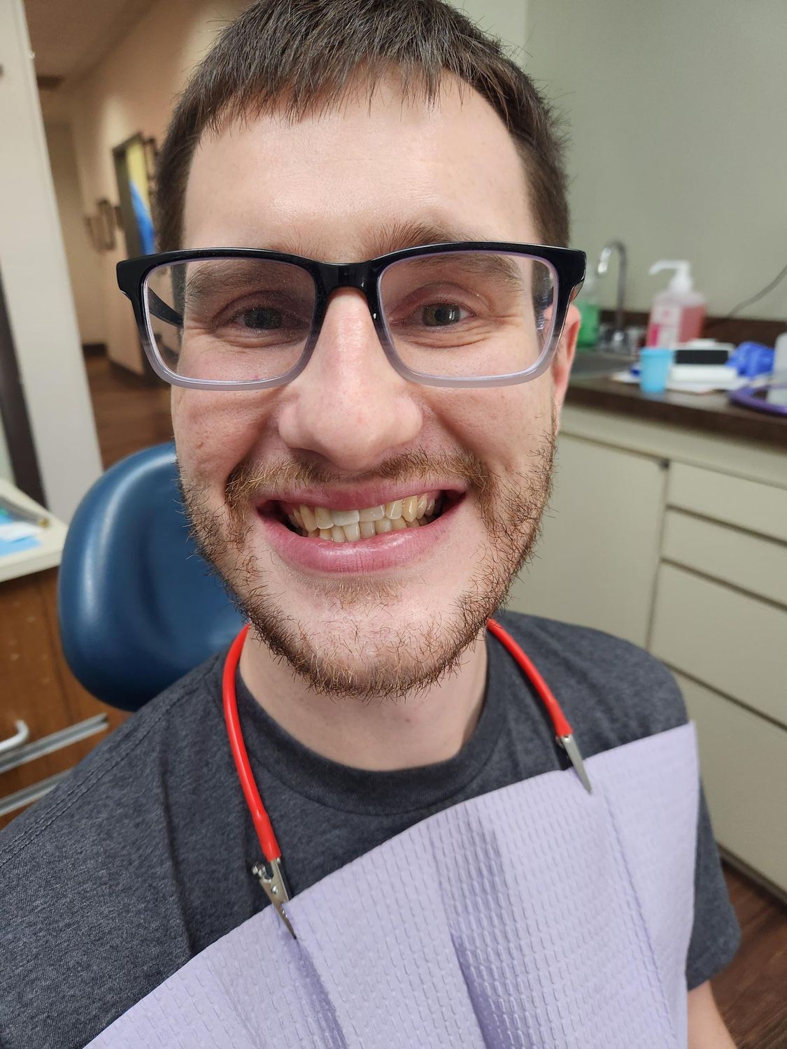Hombre con gafas en el dentista, sonriendo. Lleva un babero con cordón rojo. Habitación limpia y luminosa.