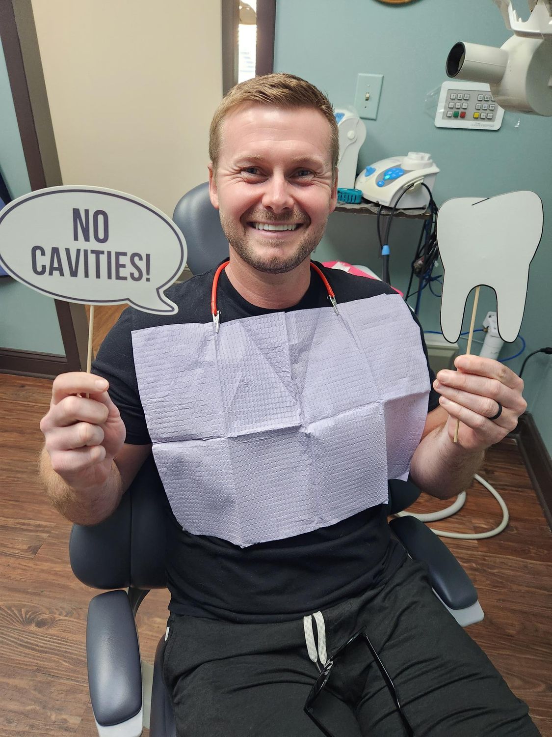 Hombre en la silla del dentista, sonriendo, sostiene un cartel de 