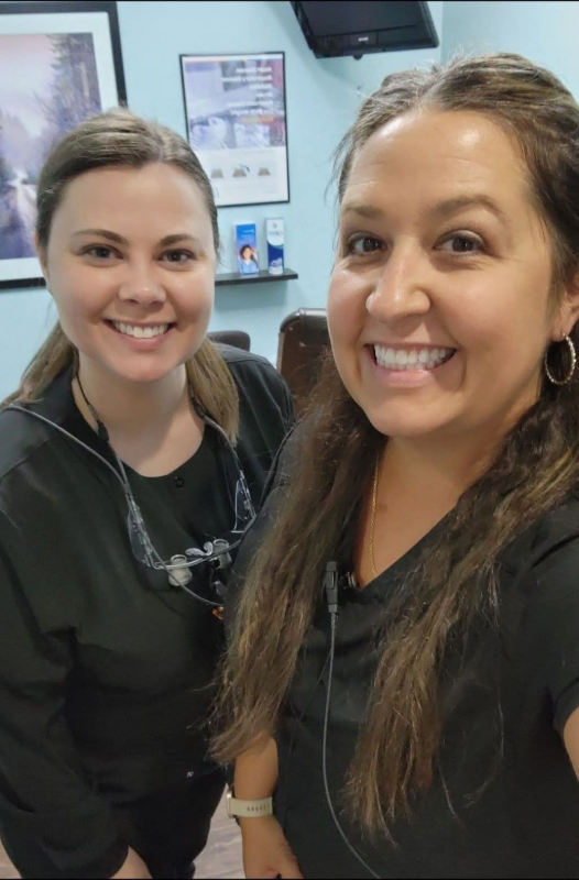 Dos mujeres sonrientes en el consultorio dental, una con cola de caballo y herramientas dentales, la otra con cabello largo.