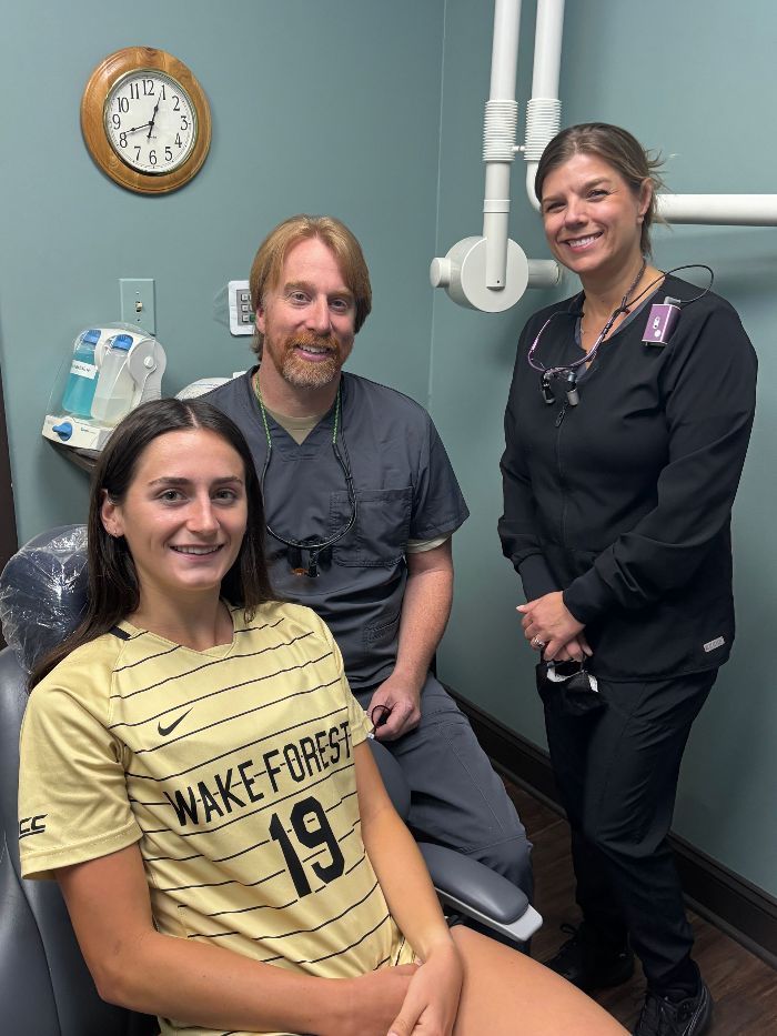 A woman in a wake forest jersey is sitting in a dental chair with two dentists.