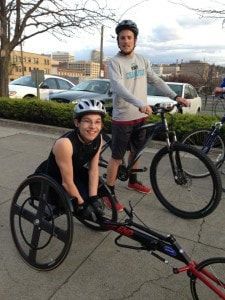 Smiling young racer in a racing wheelchair beside a cyclist on a sidewalk