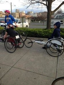 Racers in racing wheelchairs gather with a cyclist on a downtown sidewalk