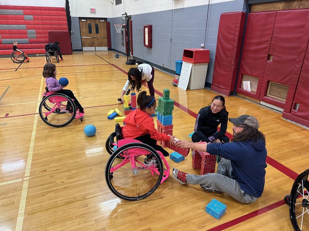 Young athletes in pink wheelchairs stack building blocks with adults