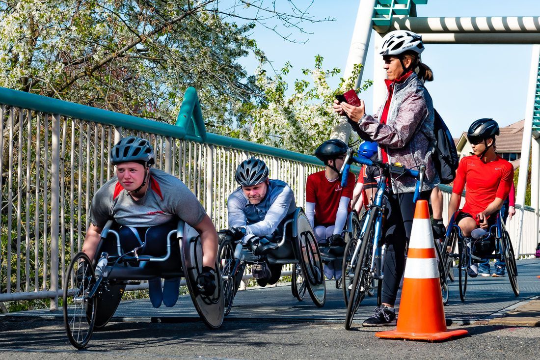 Racers line up on a footbridge as a woman with a bike checks her phone
