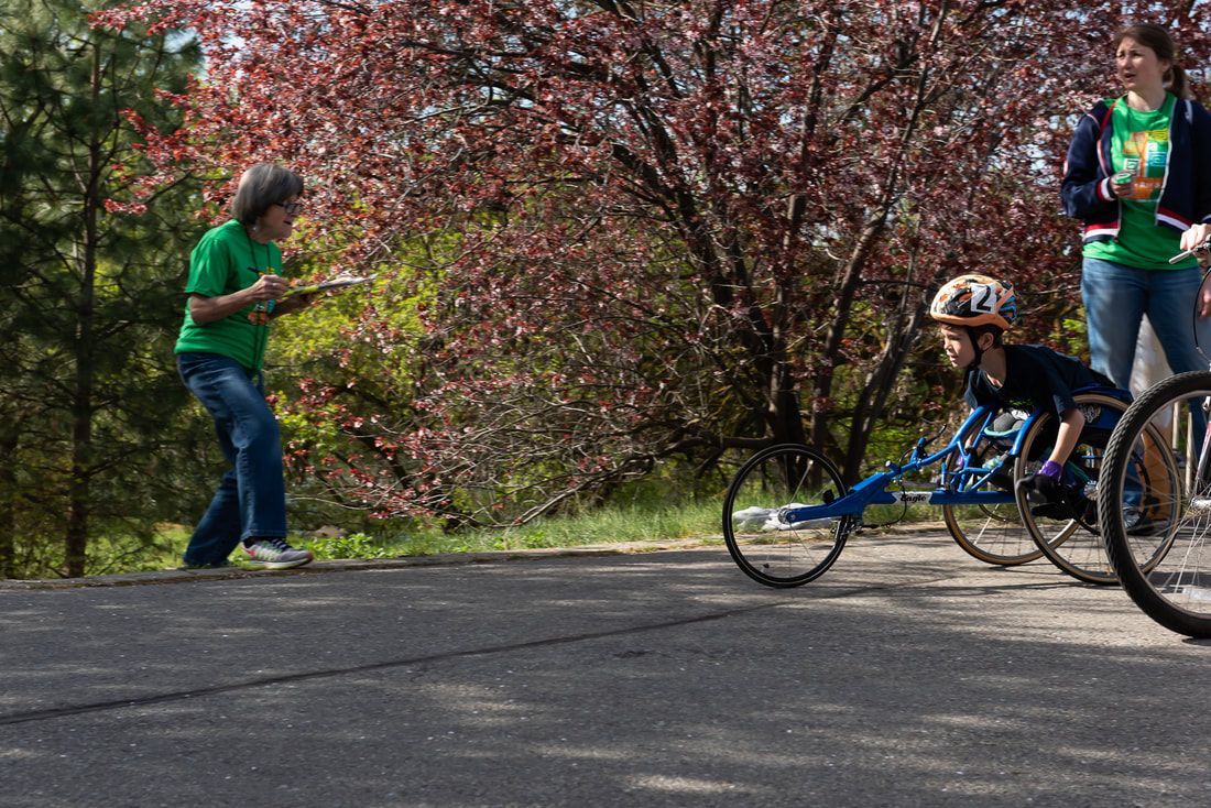 Young racer in a blue chair rolls past a volunteer in green with a clipboard