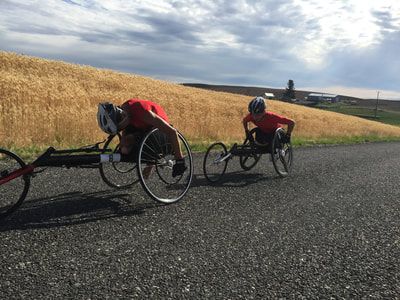 Two racers on a road below a hillside of wheat under cloudy skies