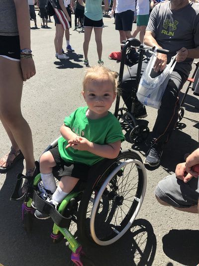 Toddler in a green shirt sits in a small sport wheelchair at an outdoor event