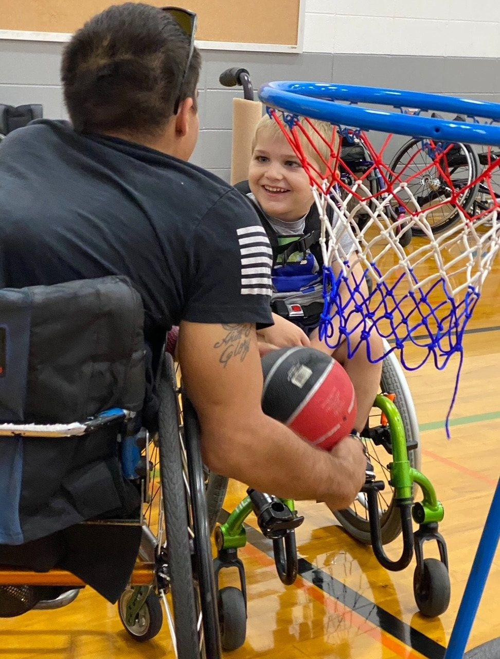 Wheelchair athlete holds a ball by a low hoop as a smiling child watches