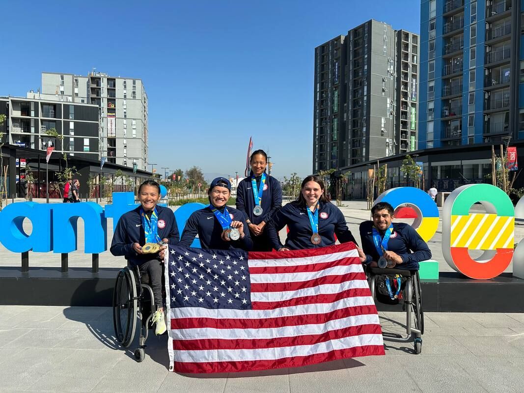 Five Team USA athletes with medals hold a US flag at the Santiago 2023 sign