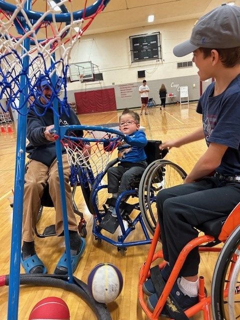Young athlete in a basketball wheelchair shooting at a lowered hoop with a coach