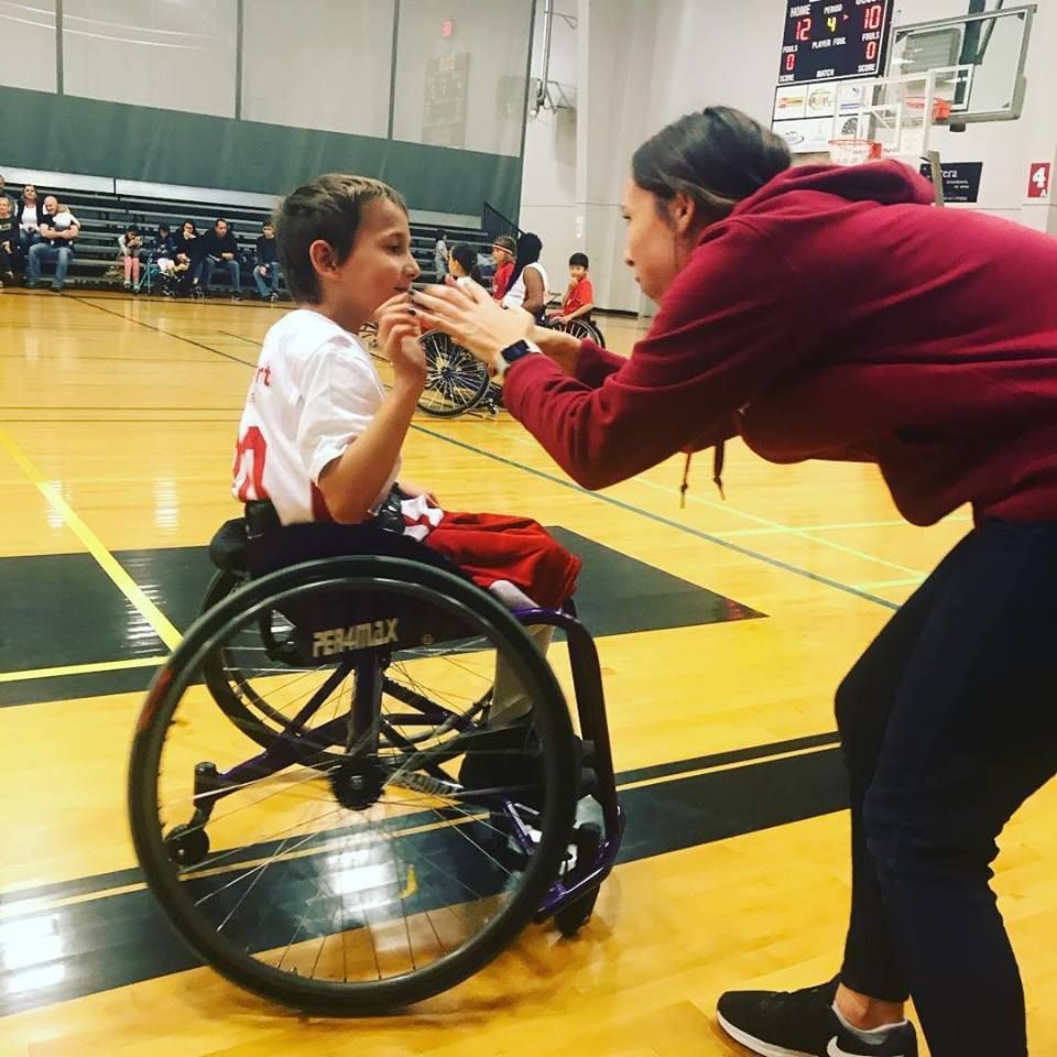 Coach in a red hoodie leans in to help a young player in a sport wheelchair