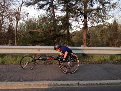 Young racer in a racing wheelchair trains along a guardrail by the trees
