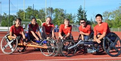 Six racers in red shirts line up in racing wheelchairs on a track