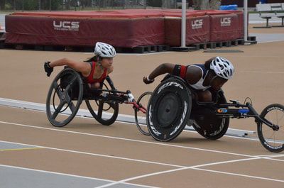 Two wheelchair racers in white helmets push side by side past red landing mats