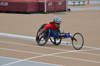 Racer in a red top and white helmet pushes a blue chair down the tan track