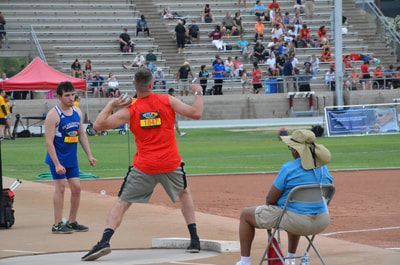 Shot putter in an orange tank throws as a seated official watches