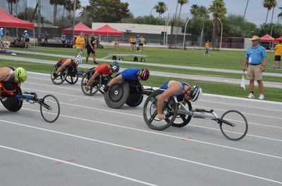 Four wheelchair racers race down the straight with palm trees behind