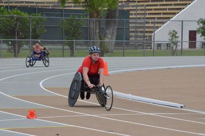 Wheelchair racer in an orange shirt leads another around the curve