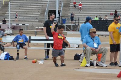 Young athlete in red holds a shot at the neck as officials sit nearby
